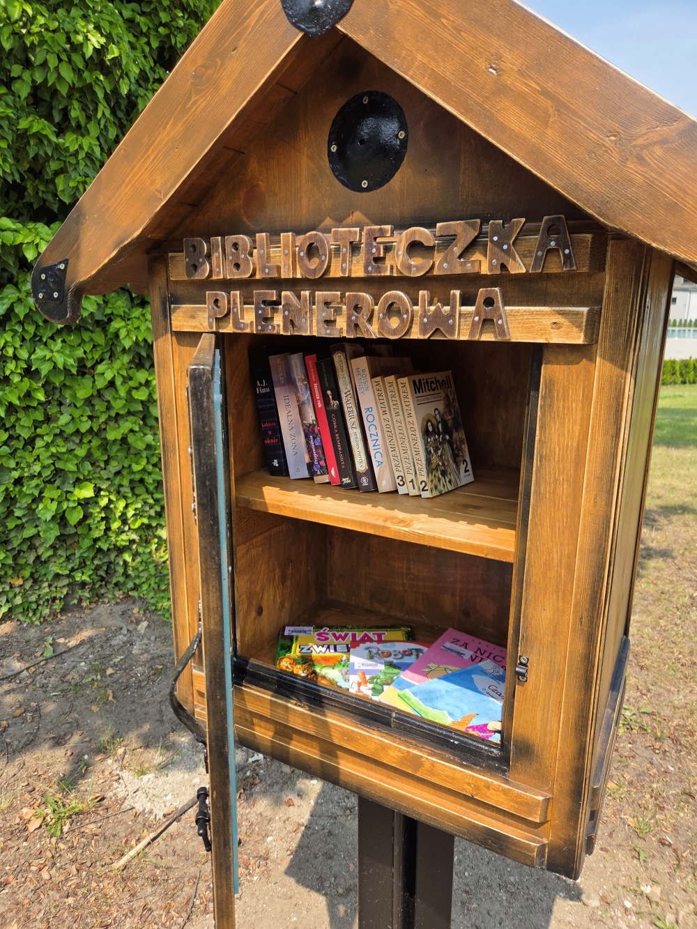 Drewniana konstrukcja z napisem "Biblioteczka Plenerowa". W środku kilka książek Drewniana konstrukcja z napisem "Biblioteczka Plenerowa". W środku kilka książek