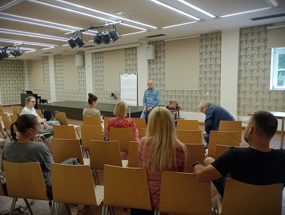 Kilkoro ludzi siedzi na krzesłach. Przed nimi stoi mężczyzna w niebieskiej koszuli