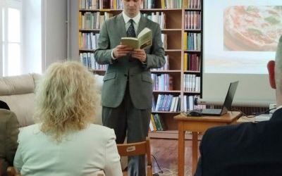 Mężczyzna w garniturze czyta książkę na głos, stojąc. Na pierwszym planie kilka osób z widowni