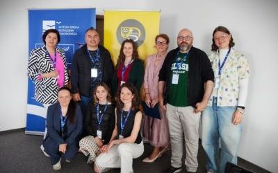 Dwóch mężczyzn i siedem kobiet pozuje do zdjęcia. Sześcioro z nich stoi, troje kuca. Wszyscy mają na szyjach zawieszone identyfikatory na niebieskich smyczach. Za grupą dwa roll-upy - jeden niebieski, drugi żółty