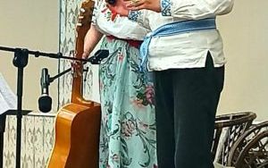 Mężczyzna i kobieta stoją na scenie. Oboje trzymają mikrofon. Kobieta w drugiej ręce trzyma gitarę. Oboje mają wyszywane koszule