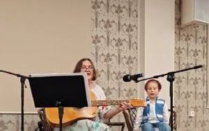 Dorosła kobieta i dziecko na scenie. Kobieta siedzi na fotelu, trzyma gitarę i patrzy na pulpit z nutami. Dziecko obok siedzi na fotelu.