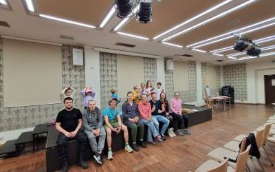 8 dorosłych osób siedzi obok siebie na scenie i pozują do zdjęcia. Za nimi siedmioro dzieci stoi, niektóre unoszą ręce nad głowę