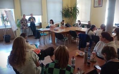 Kilkanaścioro nastolatków siedzi przy stolikach. Jedna z dziewczyn stoi i mówi do grupy