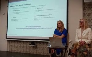 Dwie elegancko ubrane kobiety siedzą na krzesłach. Za nimi slajd prezentacji z podsumowaniem