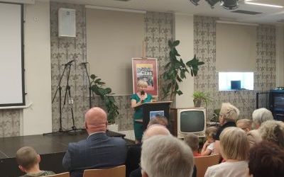 Kobieta w zielonej sukience stoi przy mównicy, trzymając mikrofon. Przed nią na krzesłach siedzi kilkoro ludzi. W tle okładka książki "Czas jak rzeka płynie" w dużym formacie