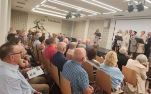 Na scenie stoi kilka osób w grupach wokół mikrofonów. Przed sceną siedzi kilkadziesiąt osób