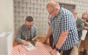 Mężczyzna w szarym garniturze podpisuje książkę. Obok stoi czytelnik