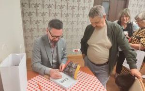 Mężczyzna w szarym garniturze podpisuje książkę. Obok stoi czytelnik