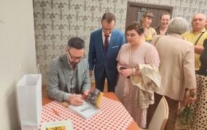 Mężczyzna w szarym garniturze podpisuje książkę. Obok stoją czytelnicy