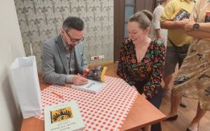 Mężczyzna w szarym garniturze podpisuje książkę. Obok siedzi czytelniczka