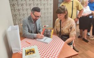 Mężczyzna w szarym garniturze podpisuje książkę. Obok siedzi czytelniczka