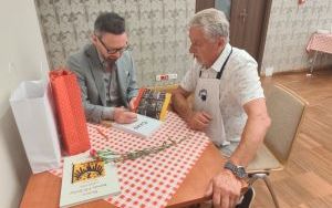 Mężczyzna w szarym garniturze podpisuje książkę. Obok siedzi czytelnik