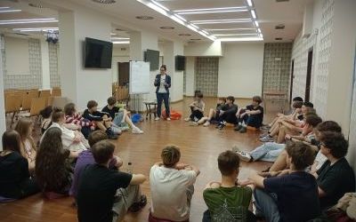 Ponad 20 osób w wieku licealnym siedzi na ziemi w kółko. Wśród nich stoi dorosła kobieta i gestykuluje