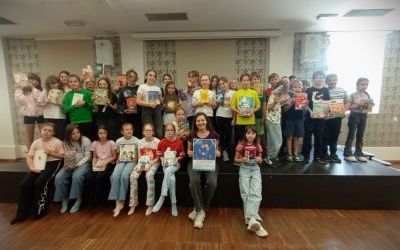 Kilkudziesięciu uczniów i jedna dorosła kobieta pozują do zdjęcia. Cześć stoi, a część siedzi na scenie. Prawie wszyscy trzymają w rękach książki