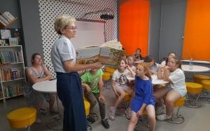 Dorosła kobieta stoi przed grupą dzieci w wieku szkolnym, które siedzą na krzesłach. Kobieta trzyma otwartą książkę