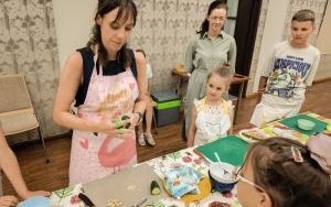 Dzieci i dwoje dorosłych stoją przy długim stole, na którym leżą zielone podkładki, talerze, sztućce i plastry arbuza
