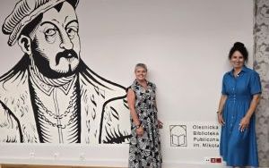 Dwie kobiety stoją przy ścianie z muralem i logo Oleśnickiej biblioteki Publicznej im. Mikołaja Reja