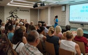 Kilkadziesiąt osób siedzi na krzesłach na sali przed stojącą kobietą