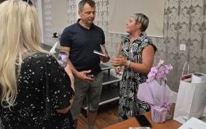 Trzy osoby stoją. Jedna robi zdjęcie, dwie pozostałe rozmawiają, obok stoi kwiat