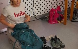 Kobieta klęczy na podłodze i trzyma plecak podróżny. Obok leżą boty trekkingowe i inne akcesoria podróżne