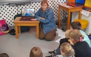 Kobieta siedzi obok stolika z laptopem, przed nią siedzi grupa dzieci