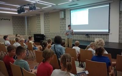 Grupa dzieci i dorosłych siedzi na krzesłach. Wyświetlana jest prezentacja