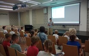Grupa dzieci i dorosłych siedzi na krzesłach. Wyświetlana jest prezentacja