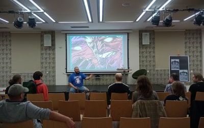 Grupa dorosłych siedzi na krzesłach. Wyświetlana jest prezentacja