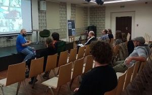 Grupa dorosłych siedzi na krzesłach. Wyświetlana jest prezentacja