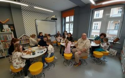 Dorośli i dzieci siedzą grupami przy okrągłych stolikach. 