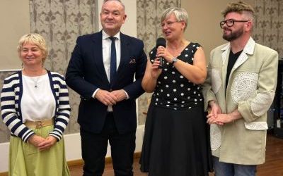 Cztery uśmiechnięte osoby stojące obok siebie w eleganckich strojach. Kobieta w środku trzyma mikrofon i przemawia.