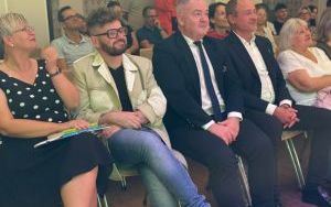 Grupa osób siedzących obok siebie w pierwszym rzędzie i patrzących w stronę sceny.