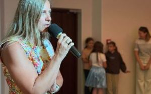 Profil długowłosej kobiety w kolorowej sukience w kwiatowe wzory, która trzyma mikrofon przy ustach.