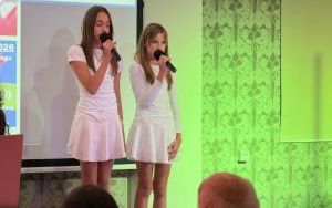 Dwie dziewczynki w białych strojach śpiewają do mikrofonów podczas występu na scenie.