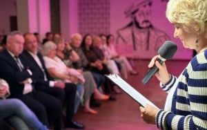 Kobieta z mikrofonem i notatkami w ręku przemawia do publiczności siedzącej w sali oświetlonej różowym światłem.