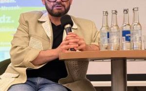 Jakub Tylman siedzi przy stoliku na scenie i mówi do mikrofonu podczas spotkania z cyklu Tydzień Szczęścia w Szkole.