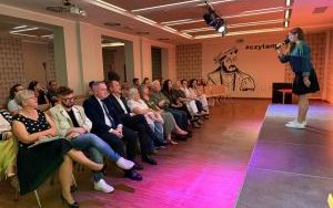 Młoda kobieta w ciemnej spódnicy śpiewa na scenie przed siedzącą na widowni publicznością w sali z portretem Mikołaja Reja.