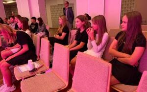 Grupa dziewcząt siedzi w rzędzie na krzesłach, uważnie oglądając program w sali oświetlonej różowym światłem.