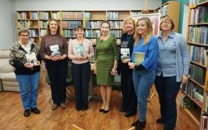 Siedem uśmiechniętych kobiet stoi w rzędzie na tle regałów z książkami w bibliotece. Kilka z nich trzyma w rękach książki.
